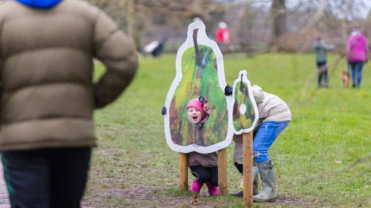 Young child peeping through large fruit cut-out on Mottisfont's outdoor The Very Hungry Caterpillar winter trail